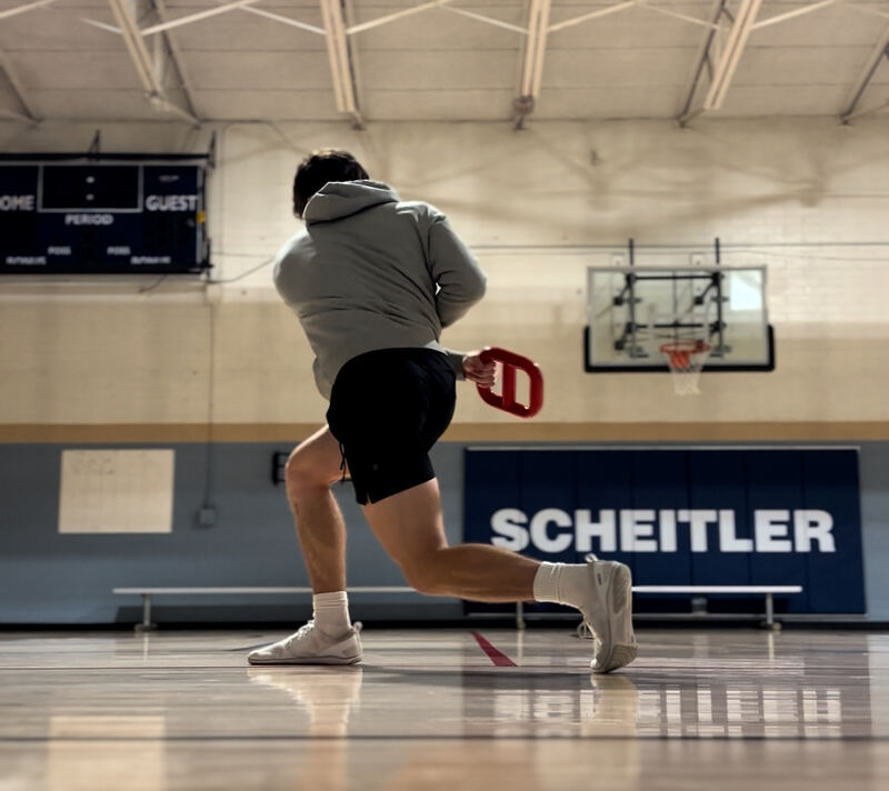 Tanner lunging across a gym floor with a red training bell held behind his back.